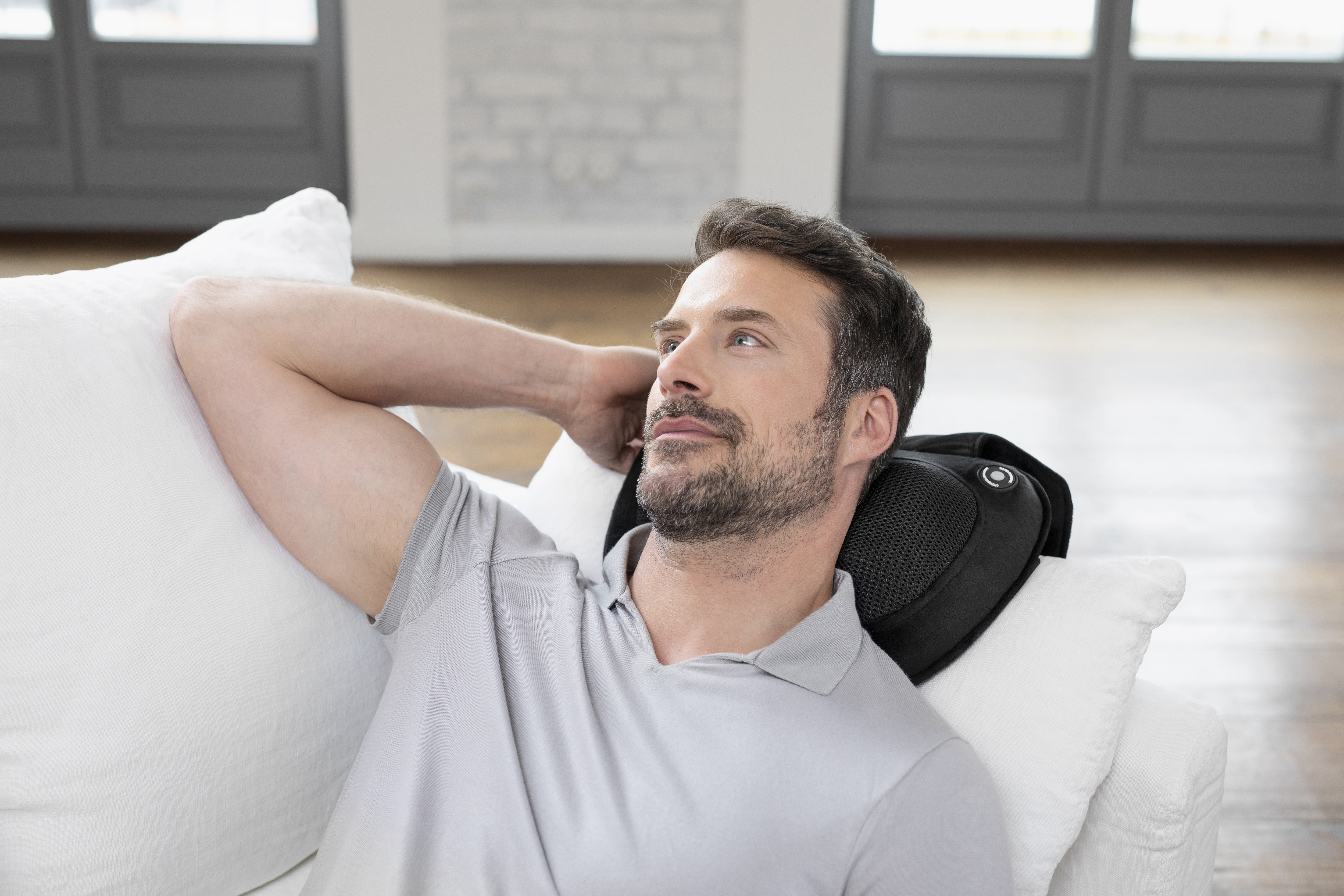 Homme portant une chemise grise utilisant un coussin de massage cervical noir sur canapé blanc.
