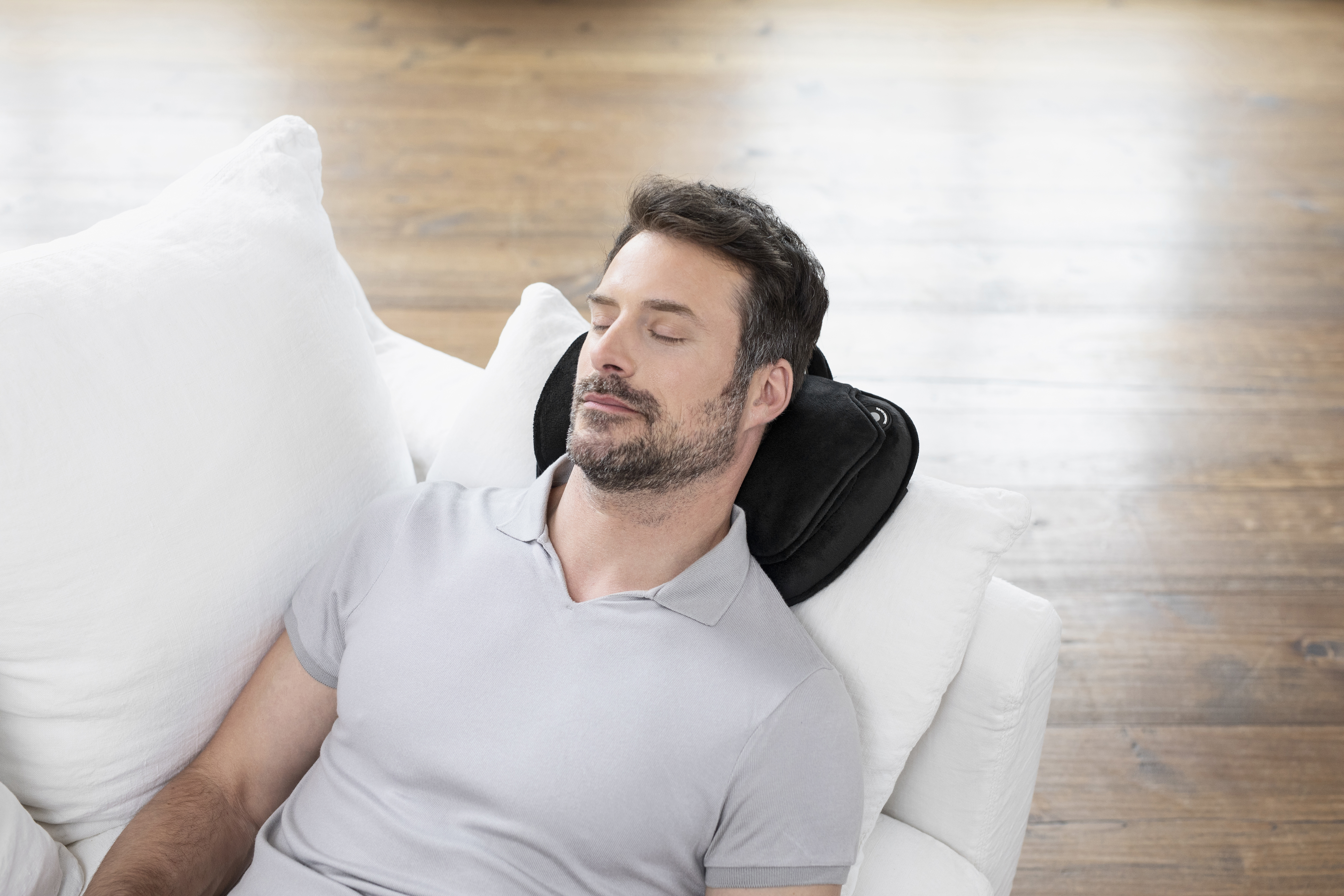 Un homme dort sur un canapé blanc avec un oreiller de massage pour le cou noir.