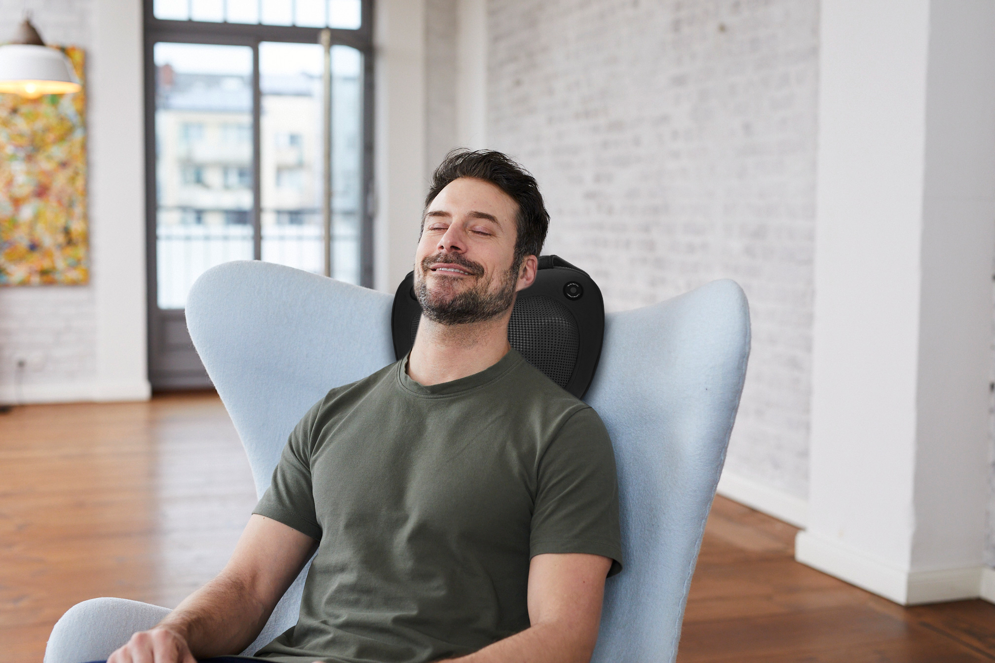 Un homme est assis dans une chaise avec un masseur de cou, souriant. Il a une barbe et les cheveux courts.