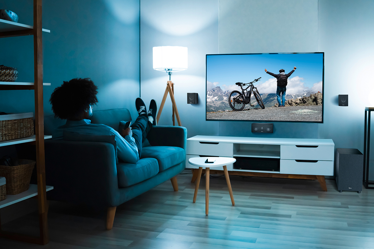 Férfi TV-t néz a kanapén hangrendszerrel és hangszórókkal. A TV hegyi képet mutat.