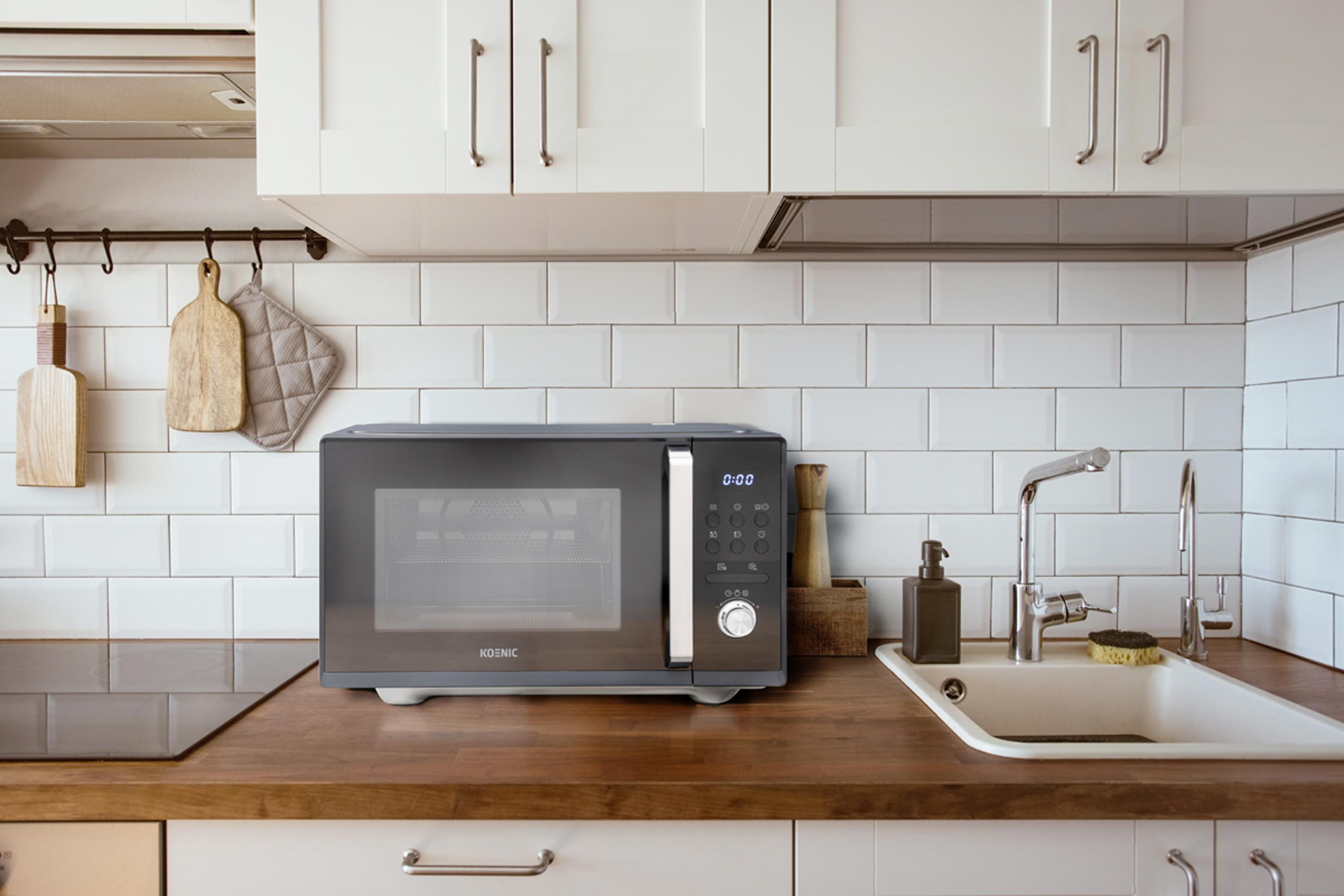 Küchenszene mit Mikrowelle auf einer Holztheke. Weiße Fliesenwand und Schränke, Spüle und Utensilien.
