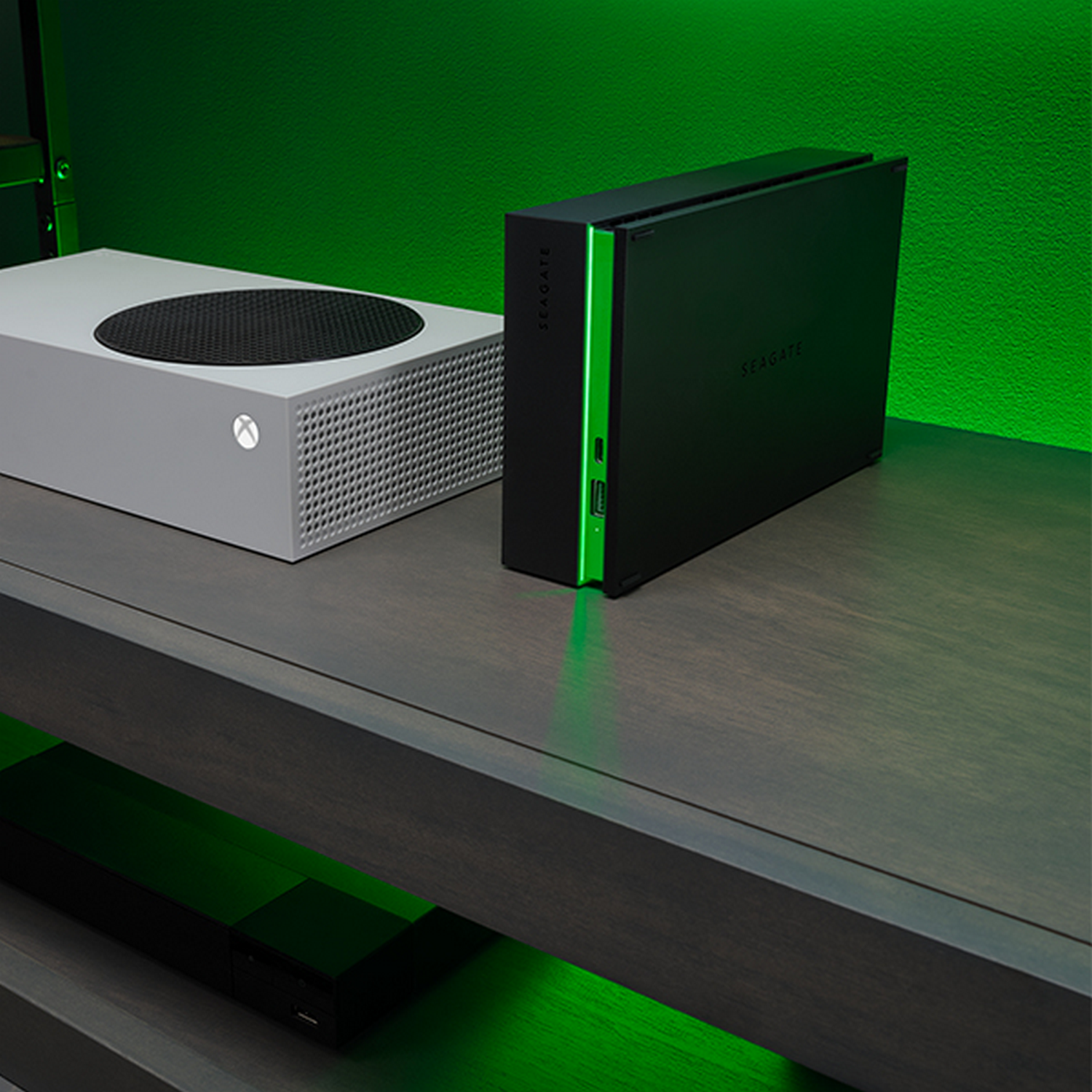 Consola Xbox blanca y disco duro externo Seagate negro sobre estante gris con luces verdes.