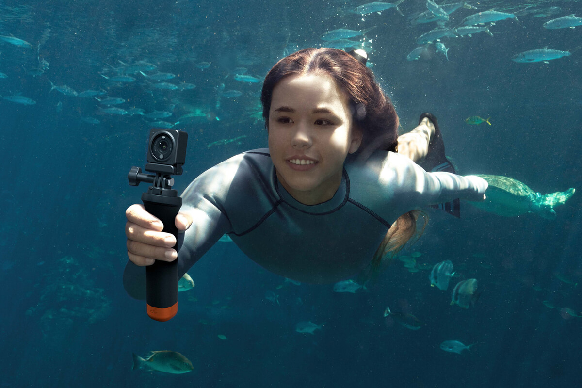 Eine Person schwimmt unter Wasser mit einer Kamera. Sie sind von Fischen umgeben. Die Person lächelt.
