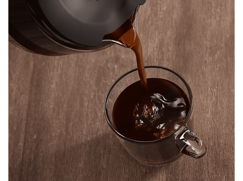 Coffee is poured into a clear glass mug from a black carafe, on a wooden table.