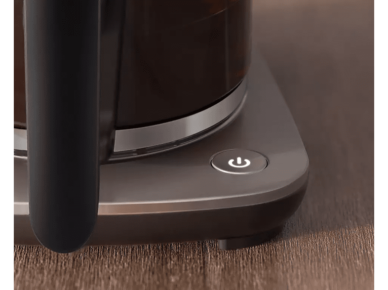 Close-up of coffee maker with power button lit. Dark glass carafe and gray base.