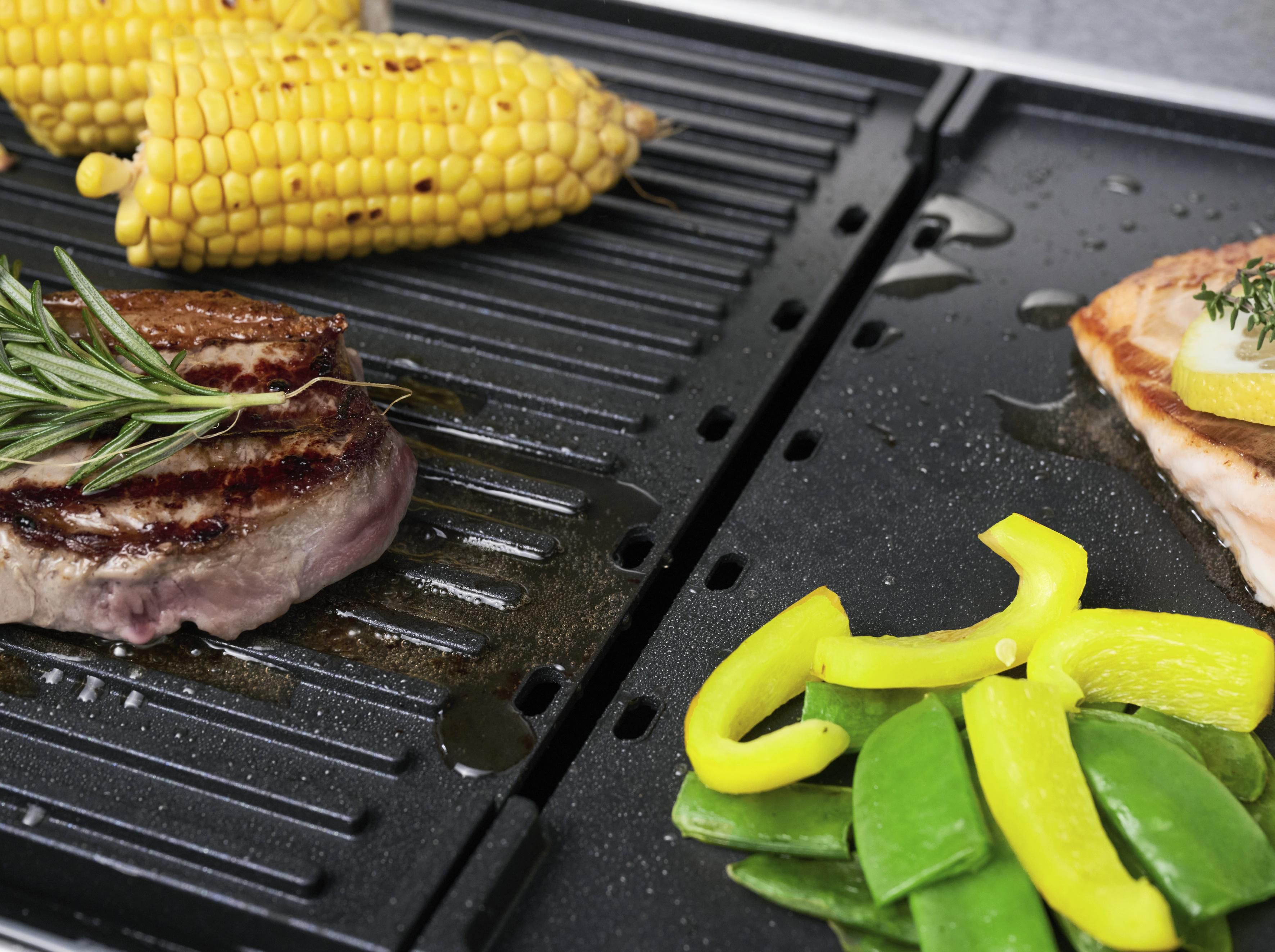 Ein Grill mit Steak, Mais und Gemüse. Das Steak hat Grillspuren. Der Mais ist gelb. Grünes Gemüse.