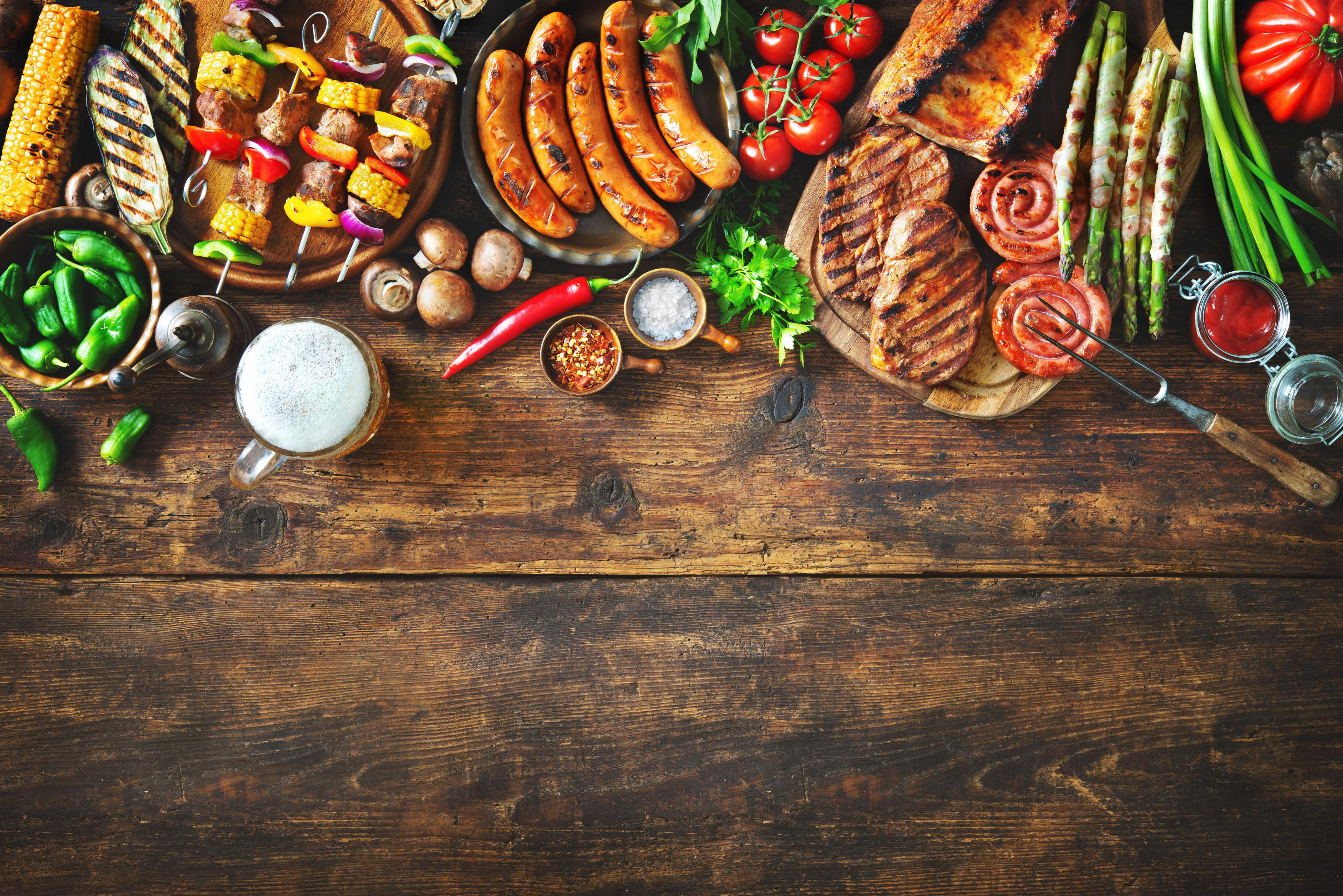 Draufsicht auf einen Holztisch mit gegrilltem Fleisch, Gemüse und Bier, bereit für ein Fest.