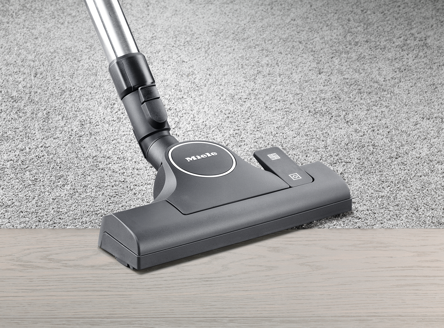 Vacuum cleaner head on a light wood floor, with a carpet in the background.
