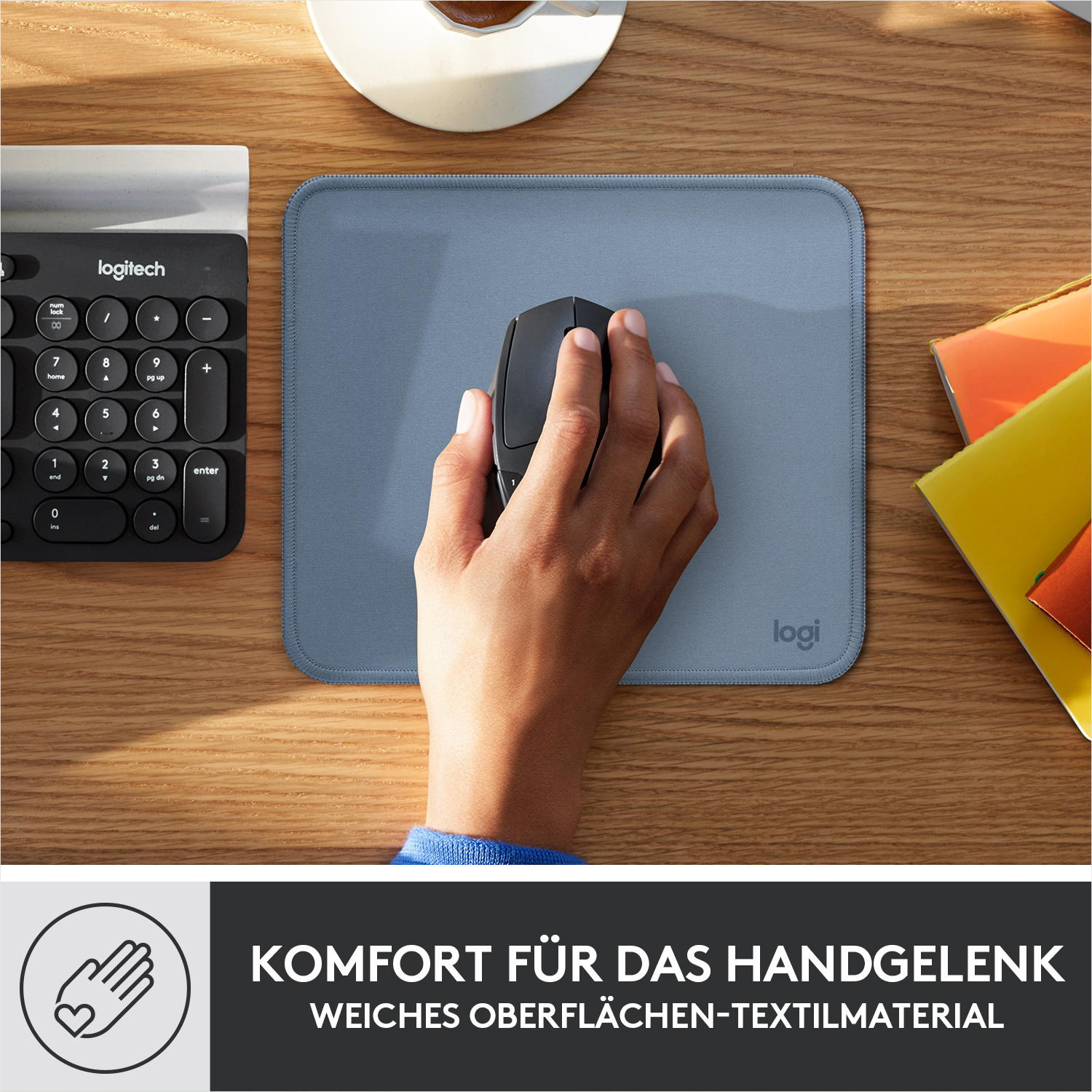 Eine Person benutzt eine schwarze Maus auf einem blauen Mauspad. Tastatur und Notizbücher auf dem Tisch.