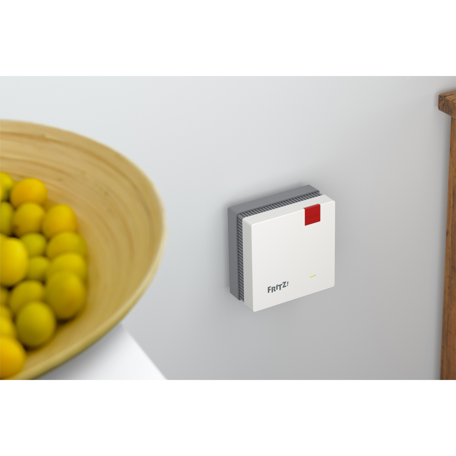 Router FRITZ!Box bianco e grigio montato su muro bianco, con ciotola di legno e frutta gialla.