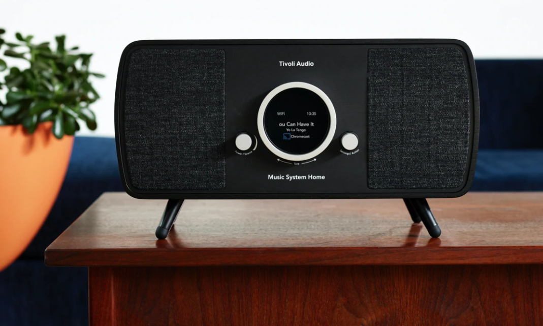 Un altoparlante nero Tivoli Audio Music System Home su un tavolo di legno. Una pianta in un vaso arancione a sinistra. Un divano blu sullo sfondo.