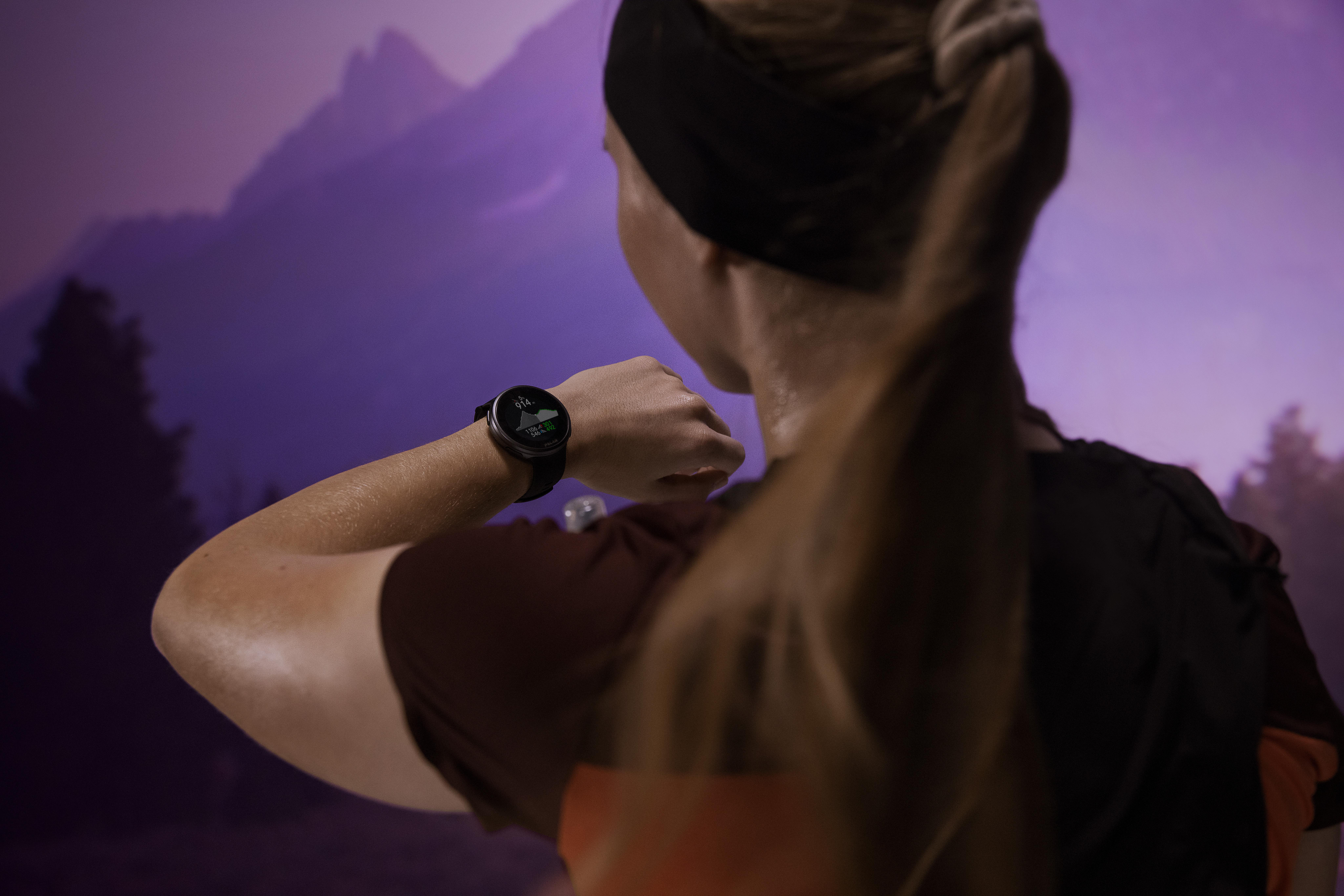 Eine Frau sieht beim Laufen auf ihre Smartwatch, Berge im Hintergrund.