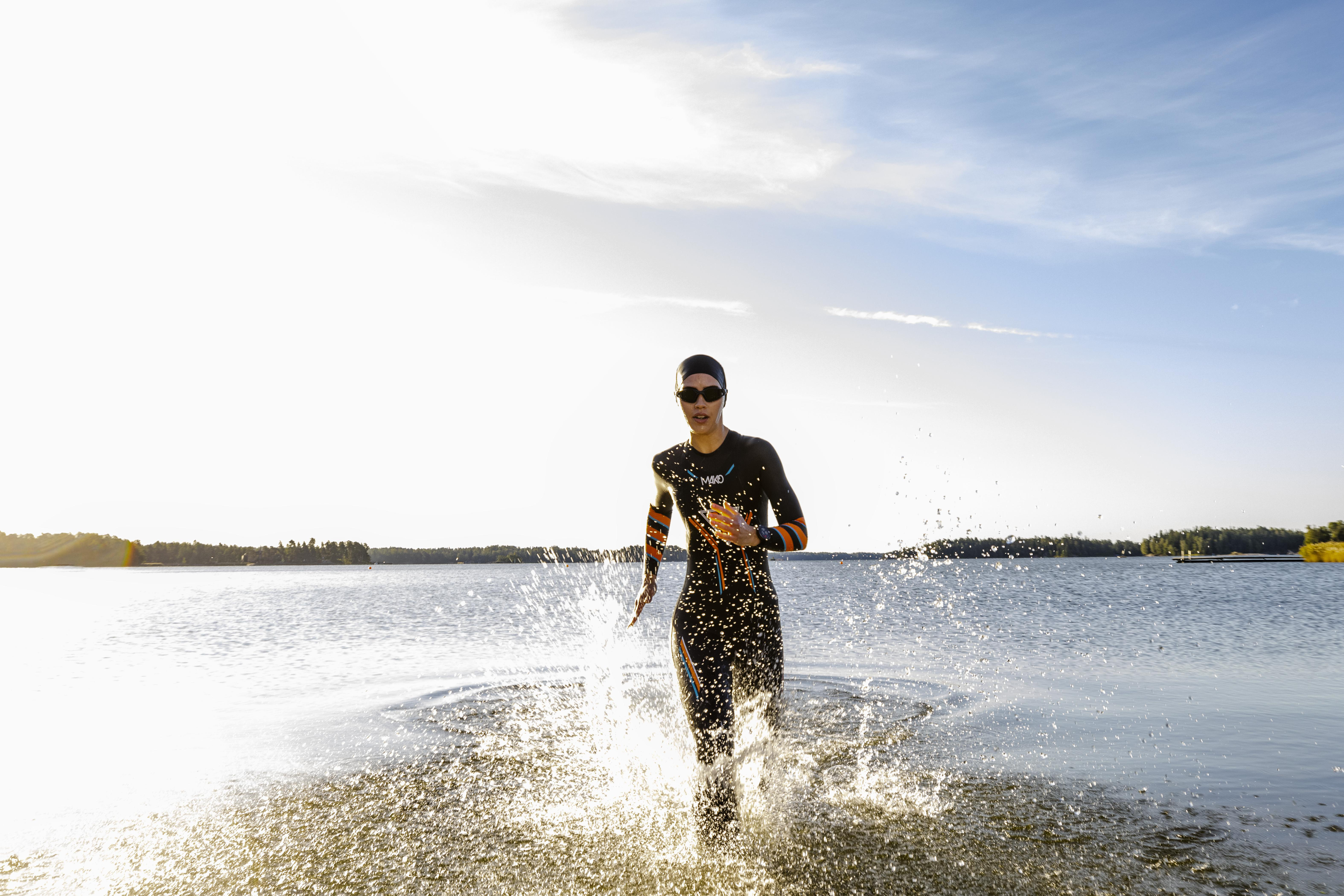 Triathlet, der aus dem Wasser rennt. Trägt Neoprenanzug, Sonnenbrille. Klares Wasser, blauer Himmel und Sonne.