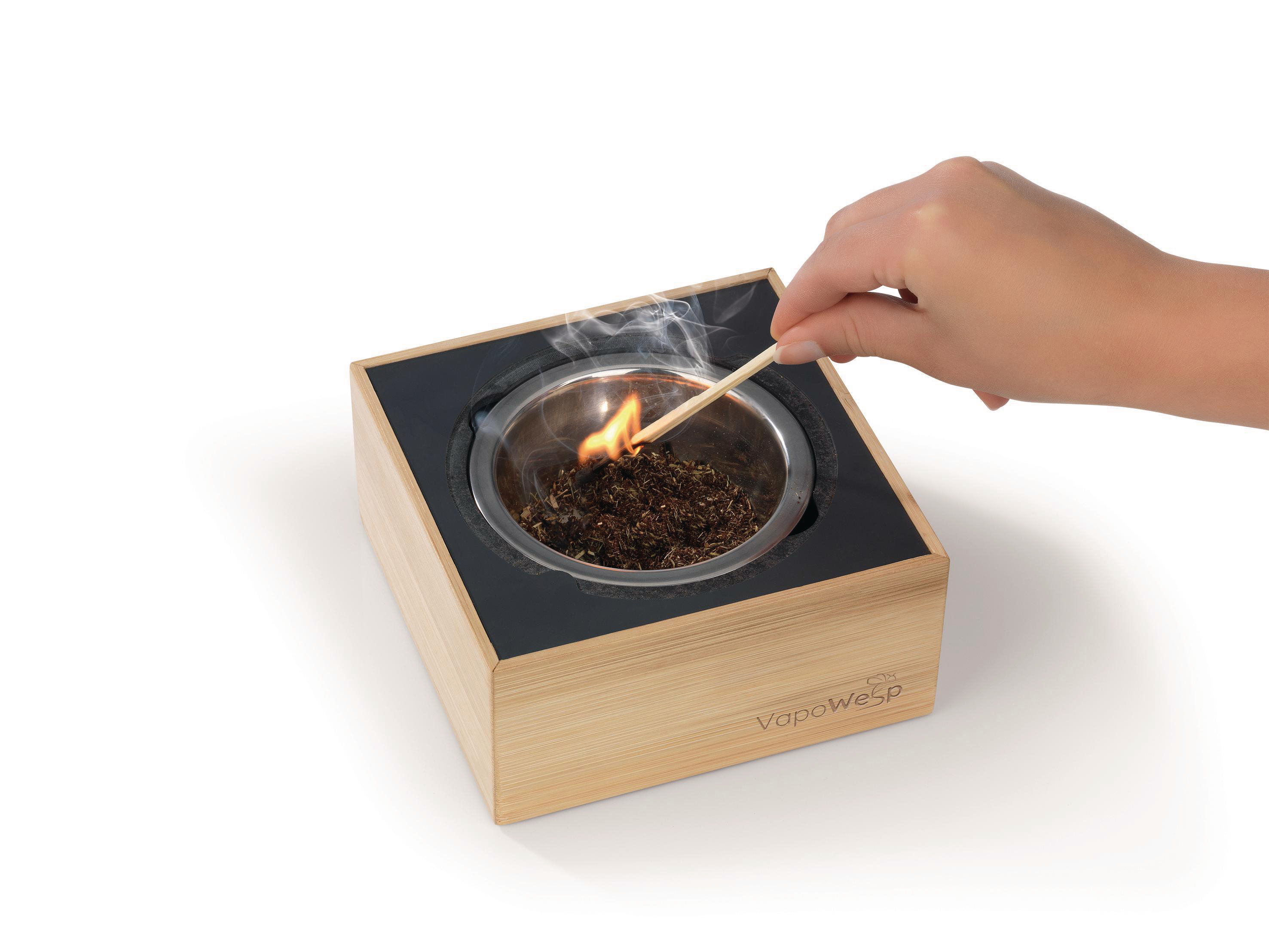Hand zündet Gerät mit Streichholz an. Gerät ist aus Holz mit Metallschale und schwarzer Oberseite.