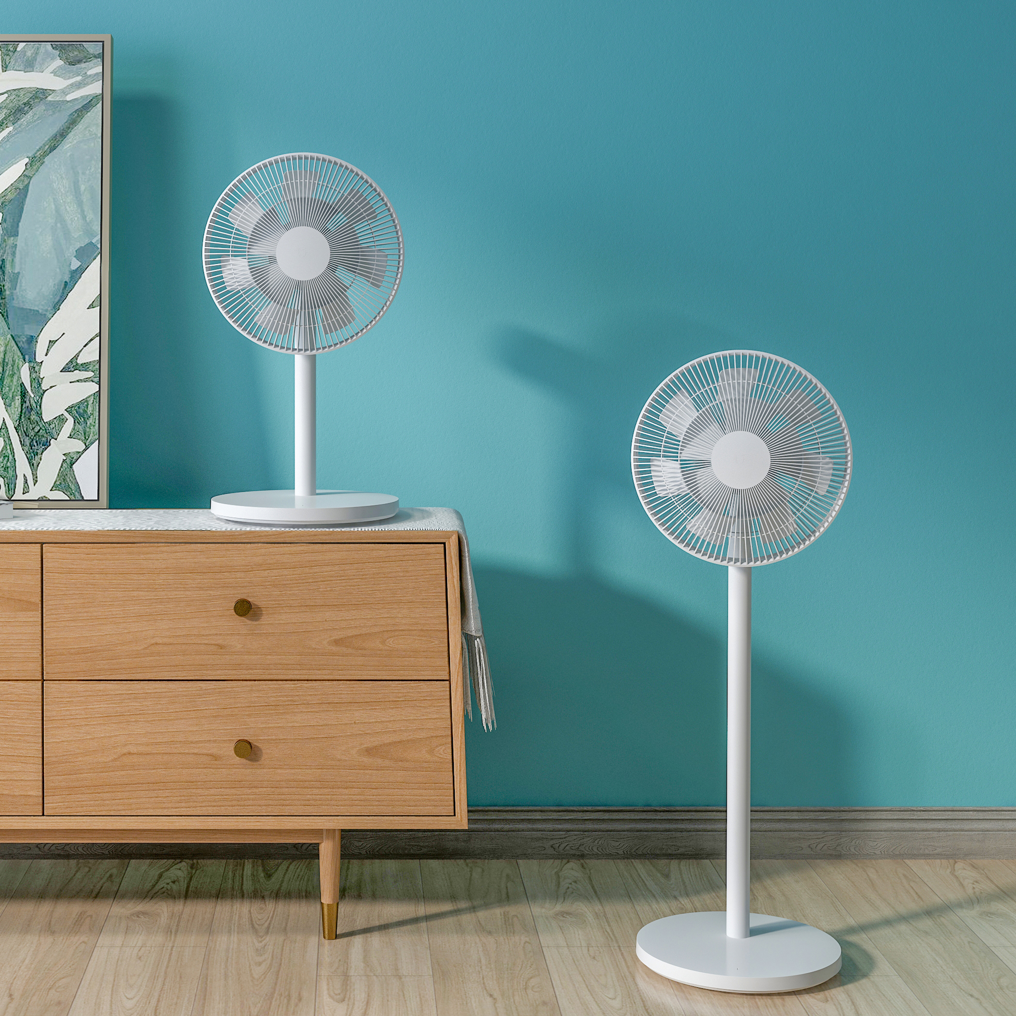 Két fehér ventilátor állványon, az egyik egy szekrényen, türkiz fal előtt.