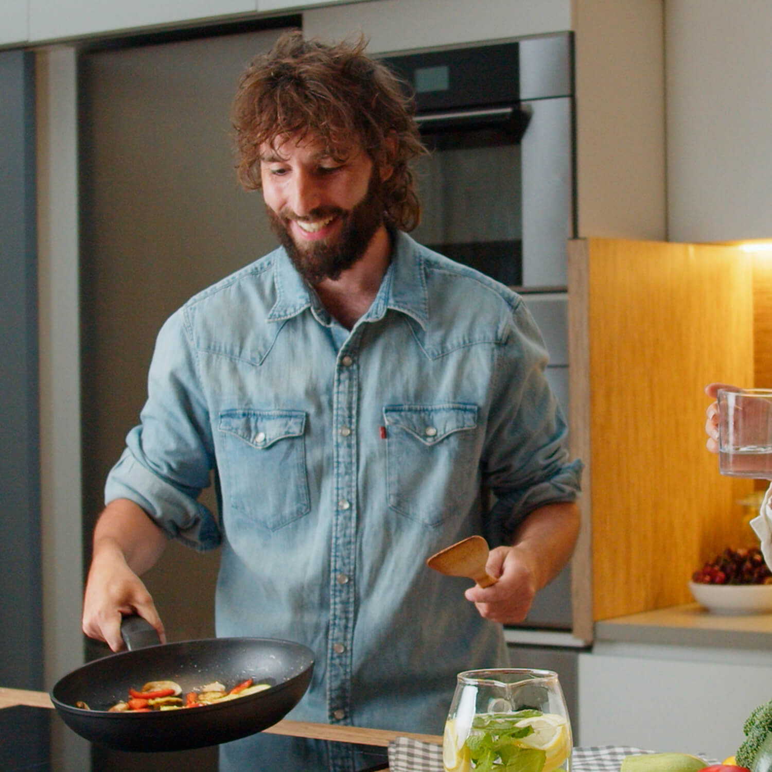 Un hombre cocinando verduras en una sartén, sonriendo, con un vaso de agua y un ambiente de cocina.