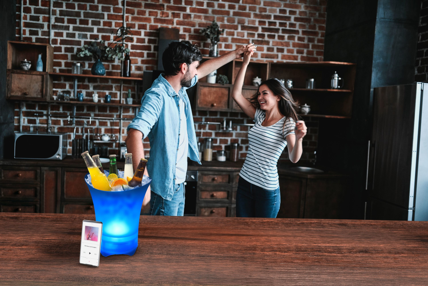 A couple dances in a kitchen with a lit ice bucket, bottles, and a phone on a wooden counter.