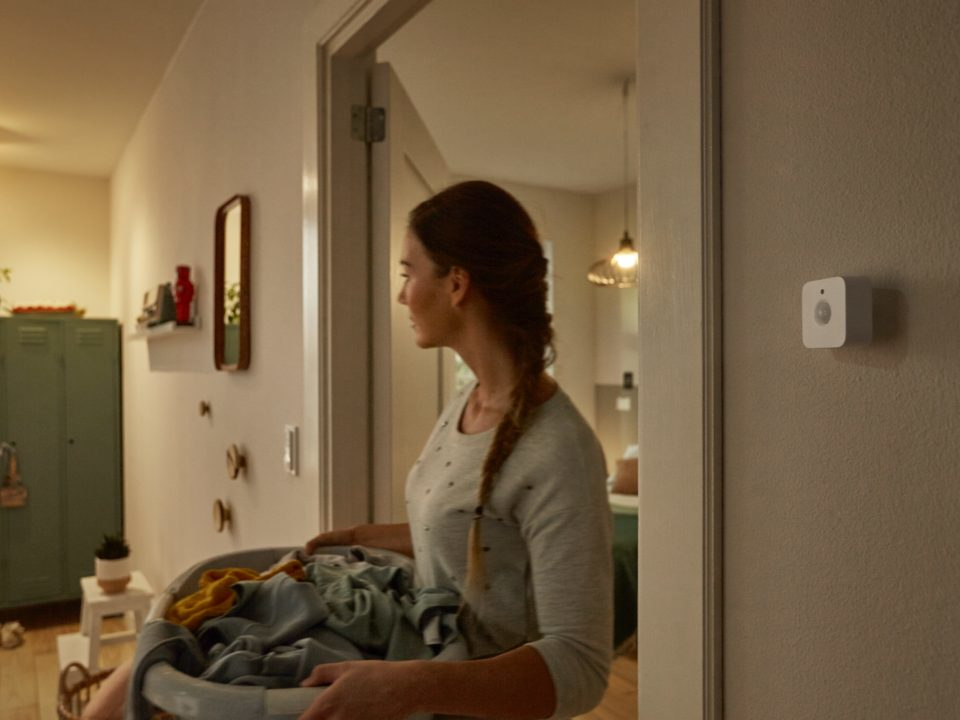 A woman with a basket of laundry enters a room with a smart home device.