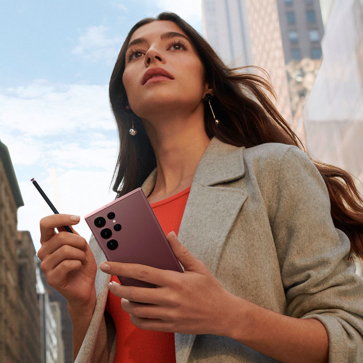 Una mujer sostiene un teléfono rosa, un bolígrafo y mira hacia arriba frente a un fondo de ciudad.