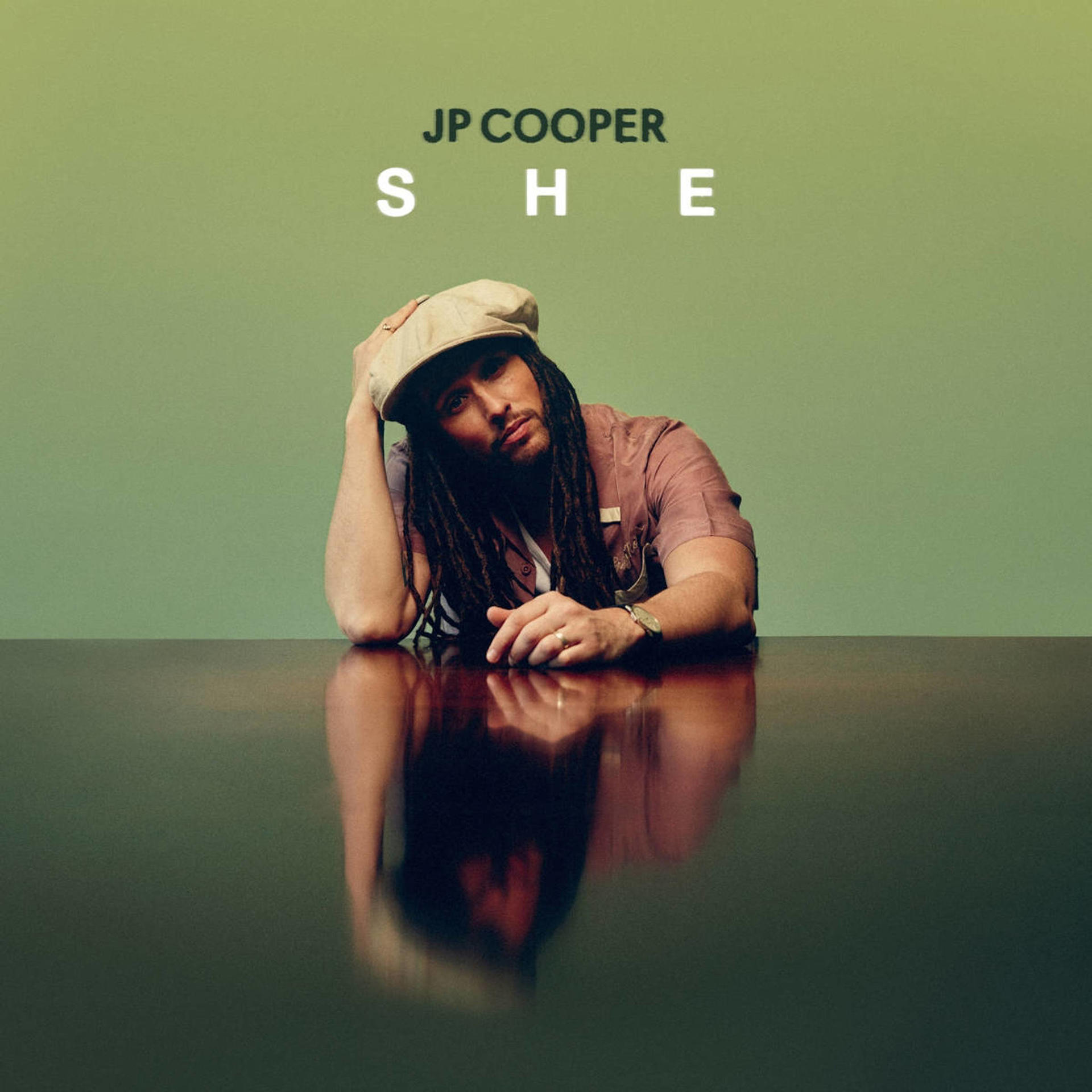 JP Cooper kalappal, egy asztalra támaszkodva, felette a SHE szóval.