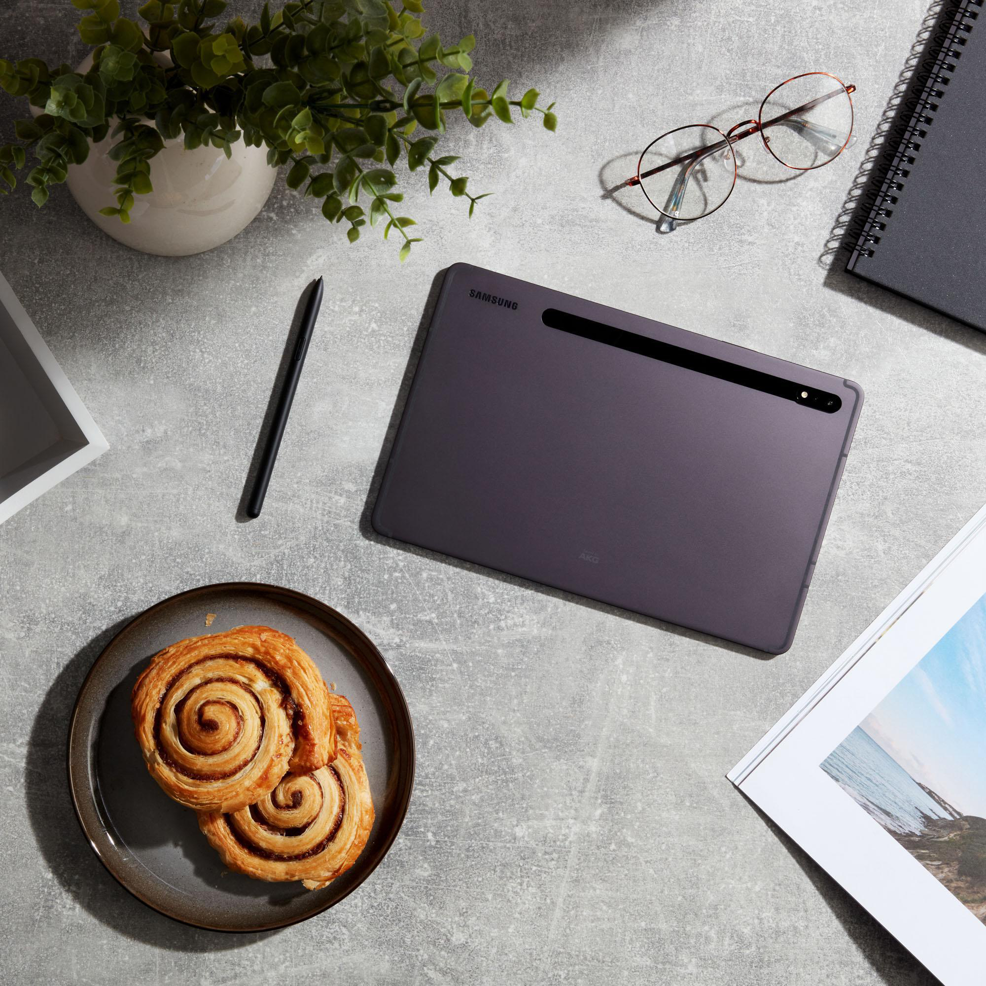 Ein Tisch mit einem Tablet, einem Stift, einem Teller mit Gebäck, einem Notizbuch, einer Brille und einer Pflanze. Die Oberfläche ist grau.