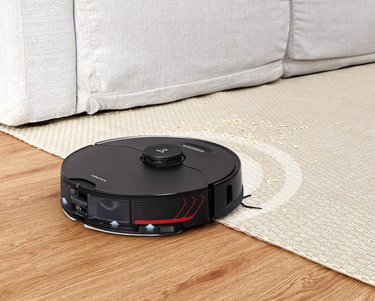 Schwarzer Roboter-Staubsauger auf Holzboden, in der Nähe eines Teppichs und eines Sofas.