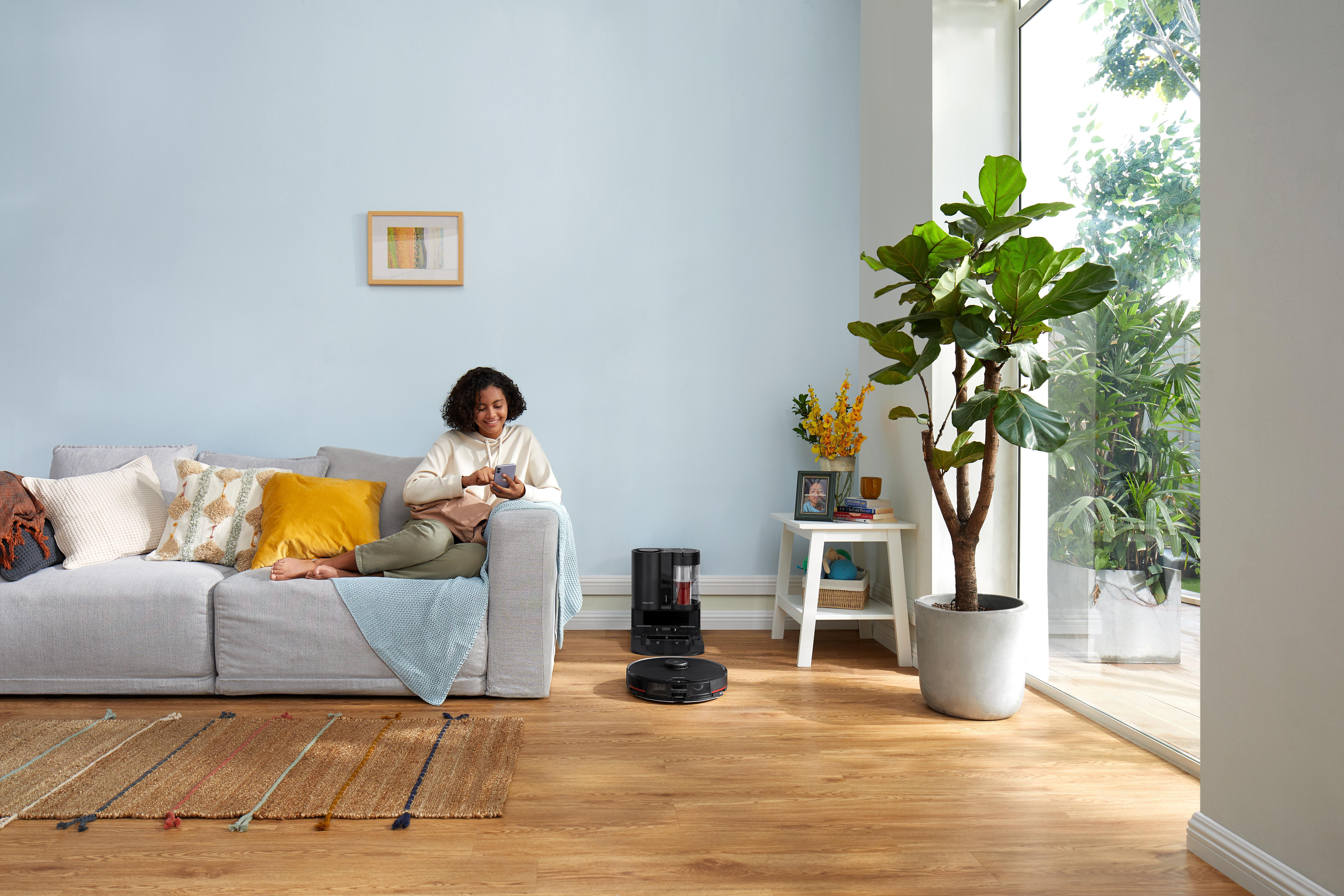 Eine Frau auf einem Sofa benutzt ein Telefon; ein Roboterstaubsauger und eine Pflanze sind im Hintergrund.