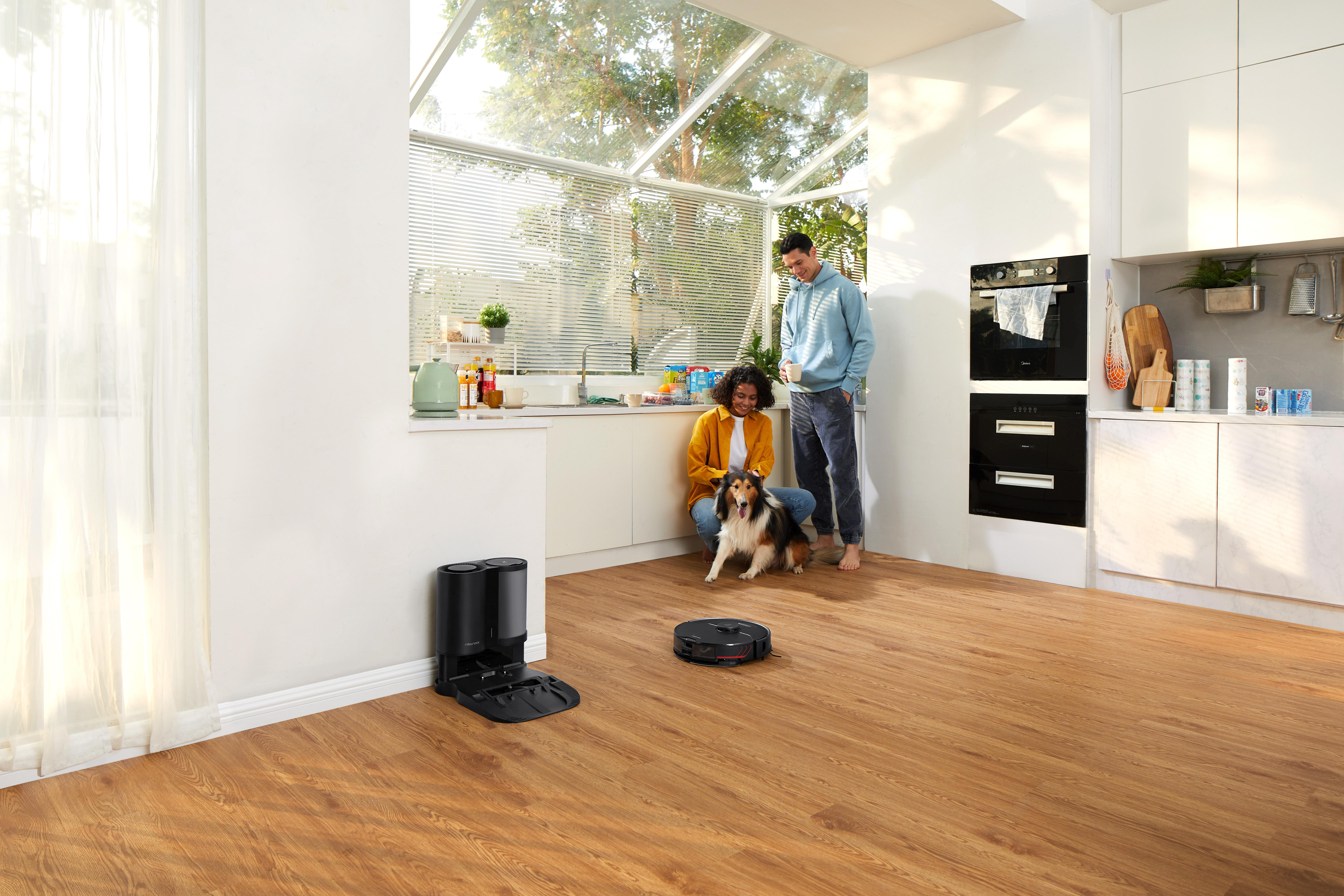 Ein Paar und ein Hund in einer hellen Küche. Ein Roboter-Staubsauger steht auf dem Boden.