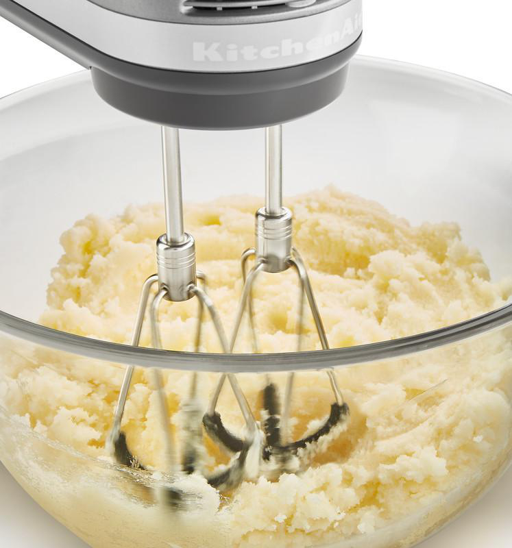 Elektrischer Mixer in einer Glasschüssel, der Kartoffelpüree mischt. Die Maschine ist silbern und grau.