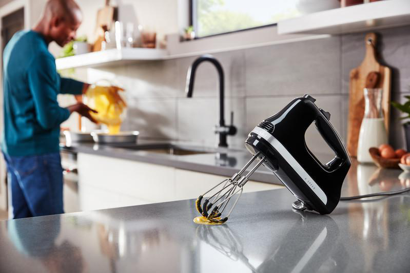 Ein Mann gießt Eier in eine Pfanne. Ein schwarzer Handmixer liegt auf der Theke mit Ei drauf.