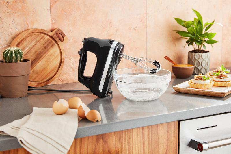 Schwarzer Handmixer neben Zutaten, Eiern und einem Schneidebrett.