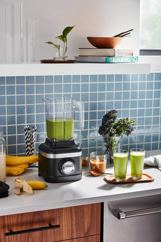 Ein schwarzer Mixer mit einem durchsichtigen Krug, gefüllt mit grüner Flüssigkeit, steht auf einer Theke mit Smoothies.