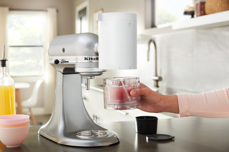 Silberner KitchenAid-Mixer mit Aufsatz. Eine Hand hält einen transparenten Behälter mit rotem Inhalt.