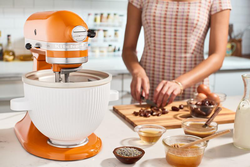 Orangener KitchenAid-Mixer mit Eiscreme-Schüssel, Zutaten und Frau, die auf einem Holzbrett hackt.