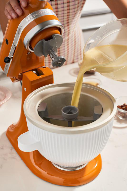 Oranger KitchenAid-Mixer. Eine Person gießt gelbe Flüssigkeit in die Schüssel.