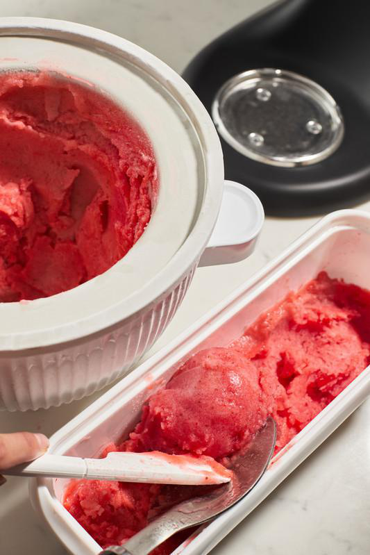 Eine weiße Eismaschine mit rosa Eis und ein Behälter mit einer Kugel Eis.