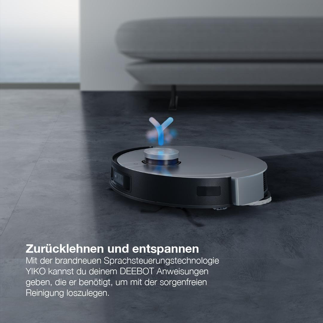 Ein silberner und schwarzer Roboter-Staubsauger auf grauem Boden, in der Nähe eines Sofas.