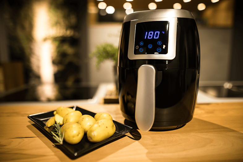 Une friteuse à air noire avec des pommes de terre sur une assiette, posée sur une table en bois. L'écran affiche la température.