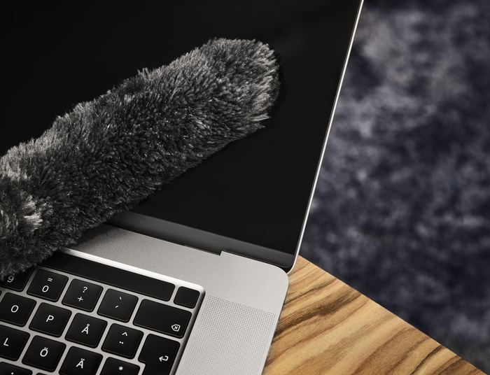 Ein Laptop wird mit einem flauschigen, grauen Staubwedel auf einer Holzoberfläche gereinigt.