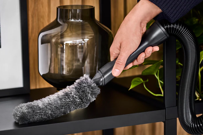 Die Hand einer Person mit einem Staubsaugeraufsatz, die ein schwarzes Regal mit einer Vase und einer Pflanze reinigt.