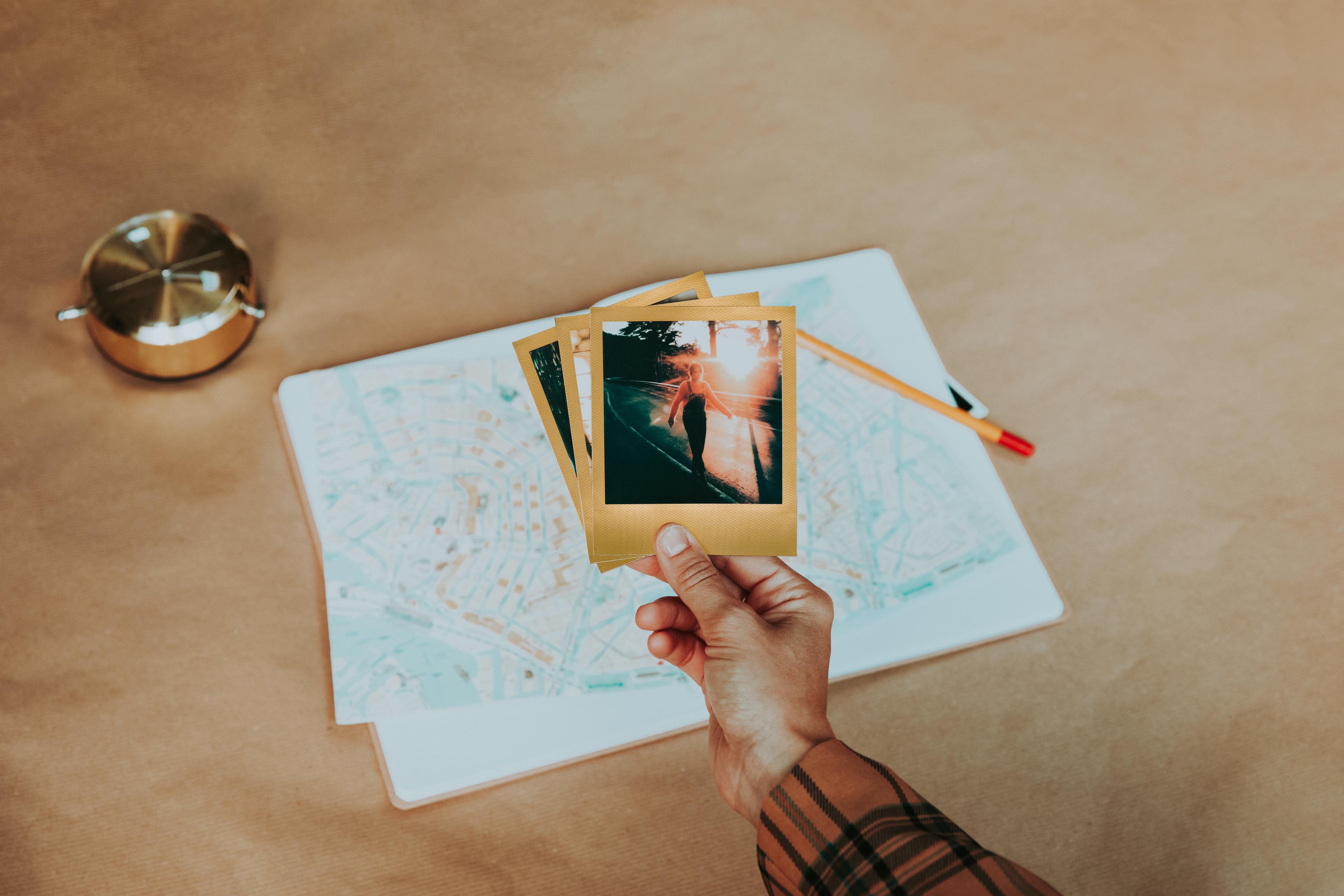 Hand hält drei Polaroid-Fotos mit einer Karte und einem Stift auf brauner Oberfläche.