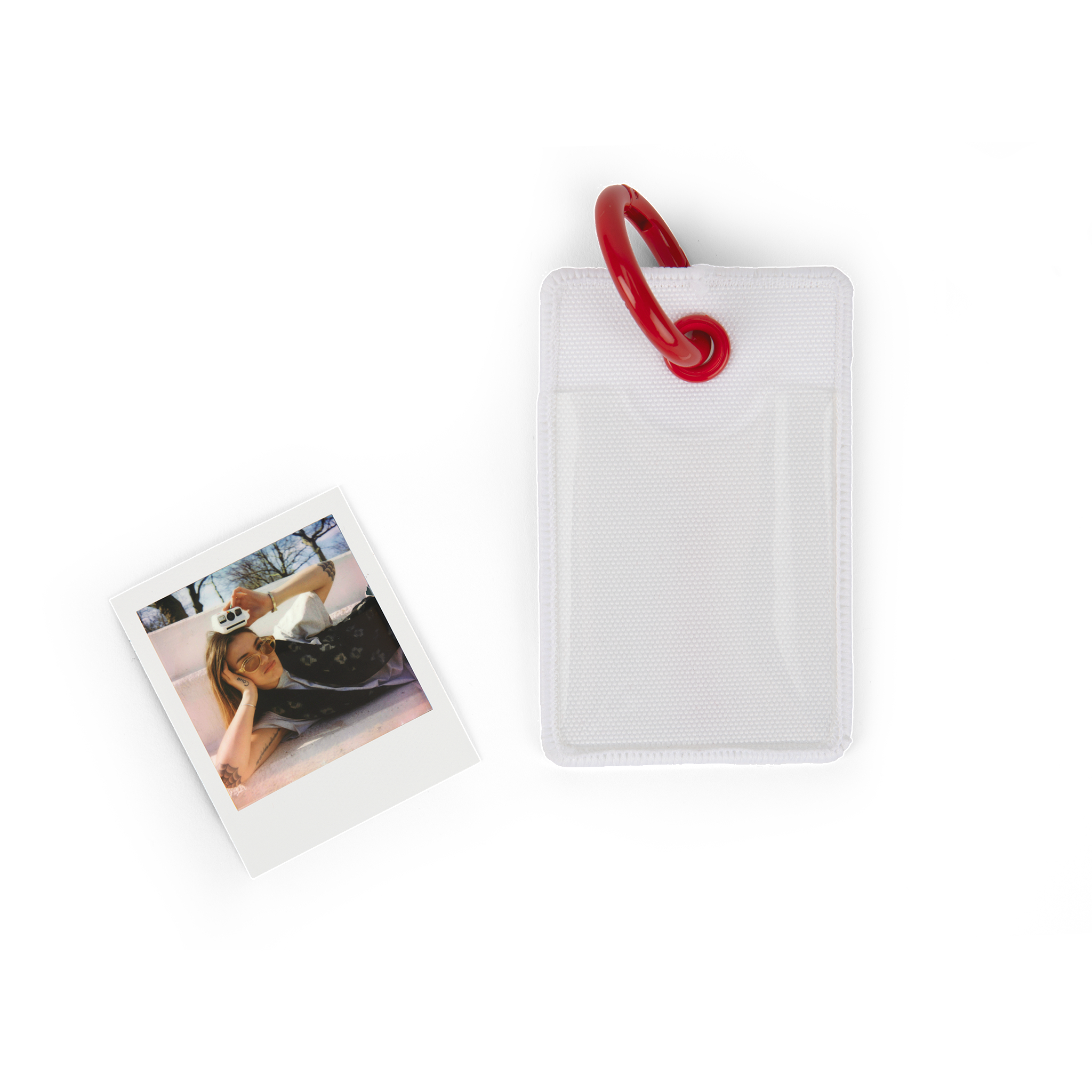 Polaroid-Foto und eine weiße Schutzhülle mit roter Schlaufe auf weißer Oberfläche.