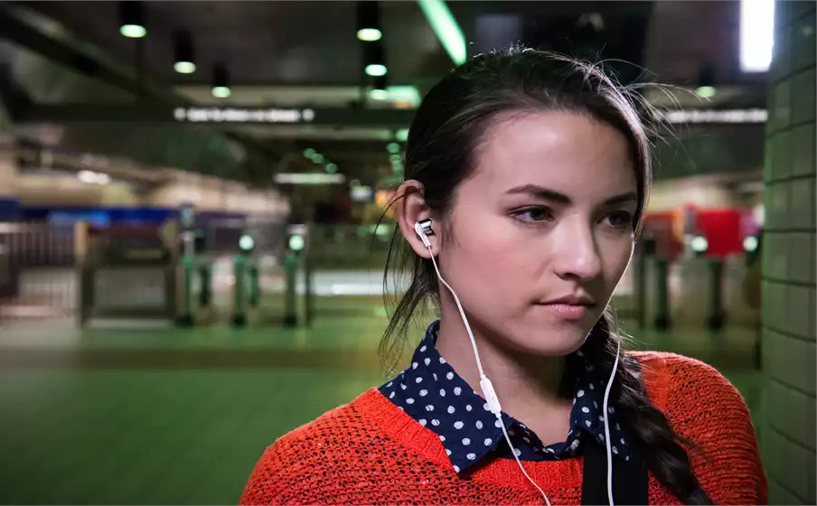 Vrouw met oortelefoons in een metrostation, draagt een rode trui en een blauw overhemd.