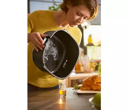 A woman pouring oil from a black air fryer into a shot glass on a wooden board.