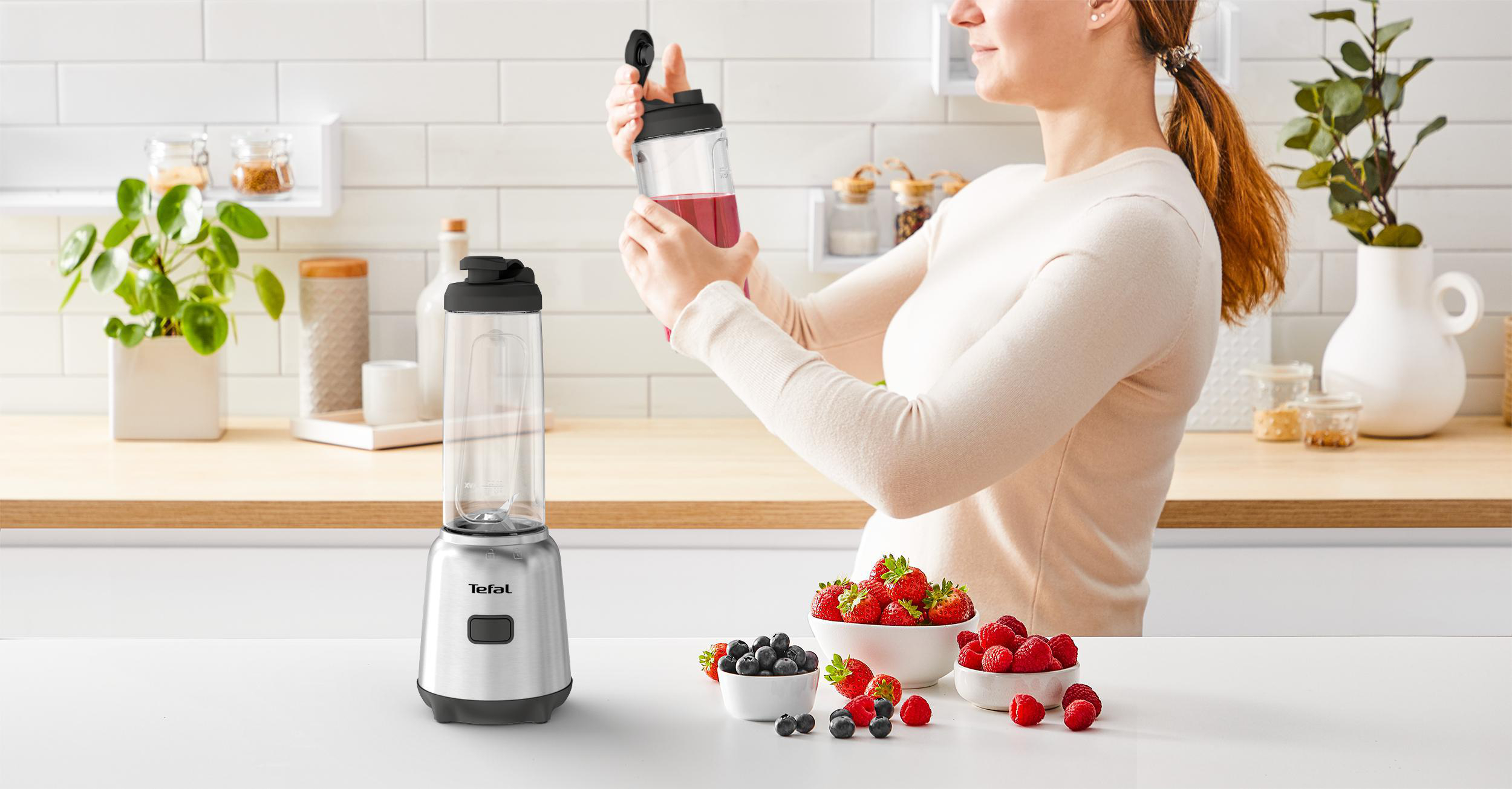 Eine Frau bereitet einen Smoothie zu und hält eine Mixbecher. Beeren und ein Mixer stehen auf einem Tisch.