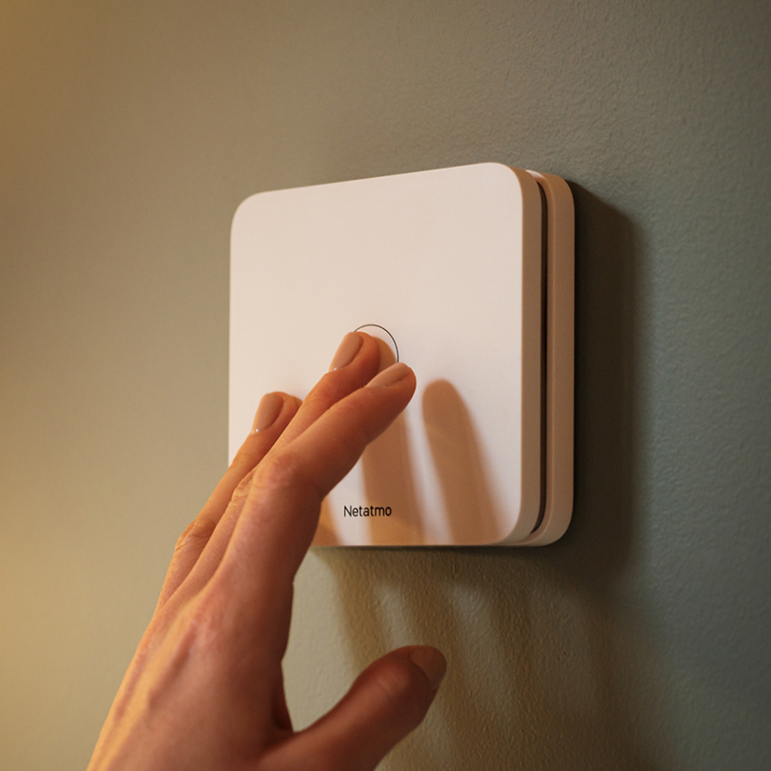 Mano presionando botón blanco Netatmo en pared verde. Diseño cuadrado. Aspecto moderno.