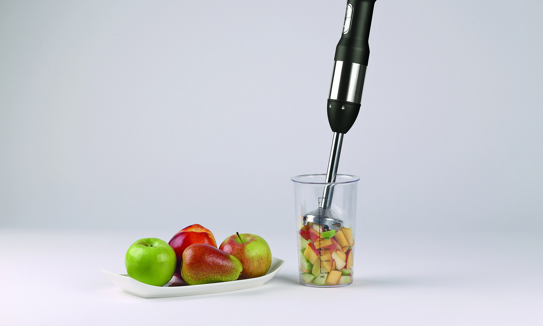 Mixeur plongeant dans un verre avec des fruits et des pommes sur une assiette.