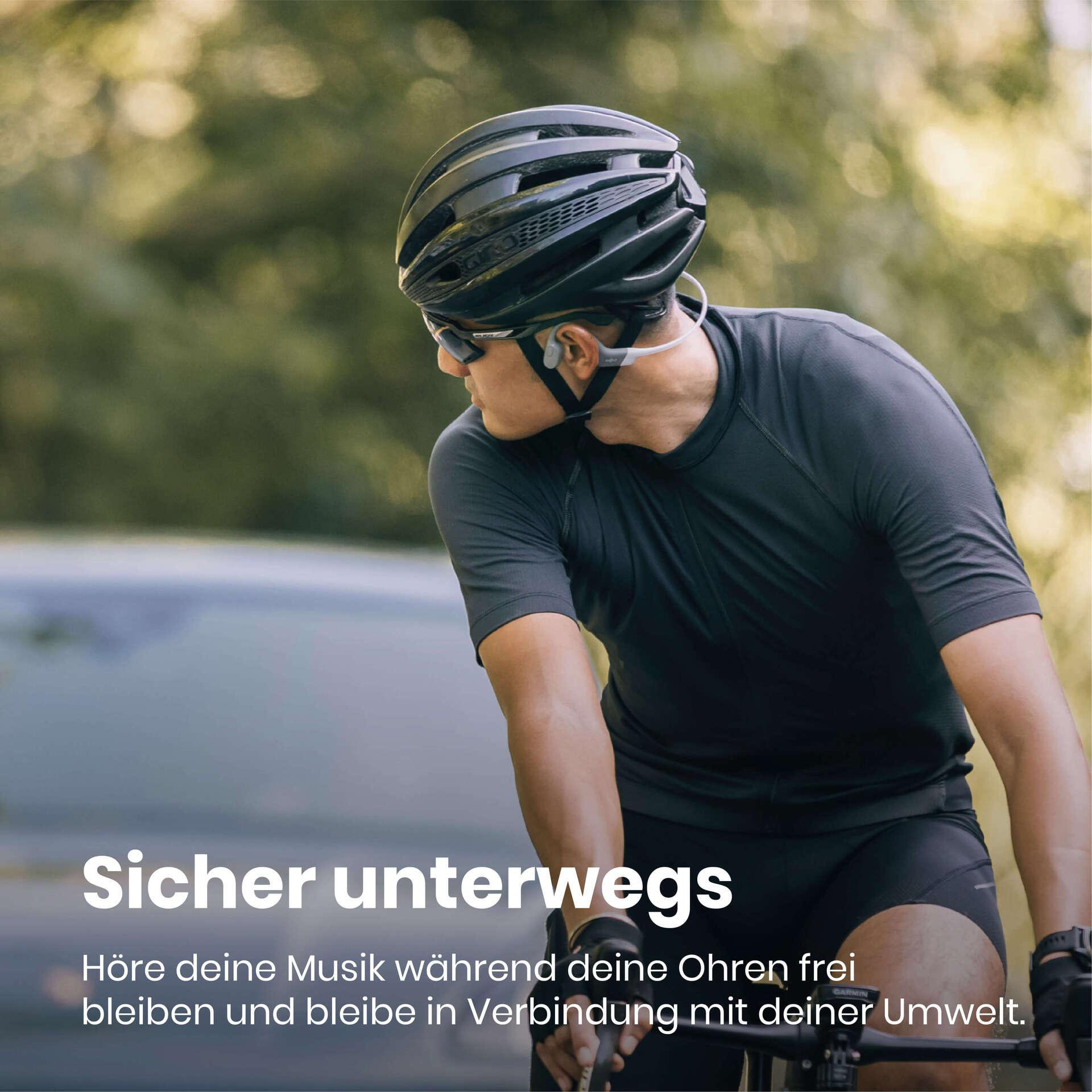 Radfahrer mit Helm und Sonnenbrille, hört Musik. Verschwommener Hintergrund mit Bäumen und Auto. Text: Sicher unterwegs.