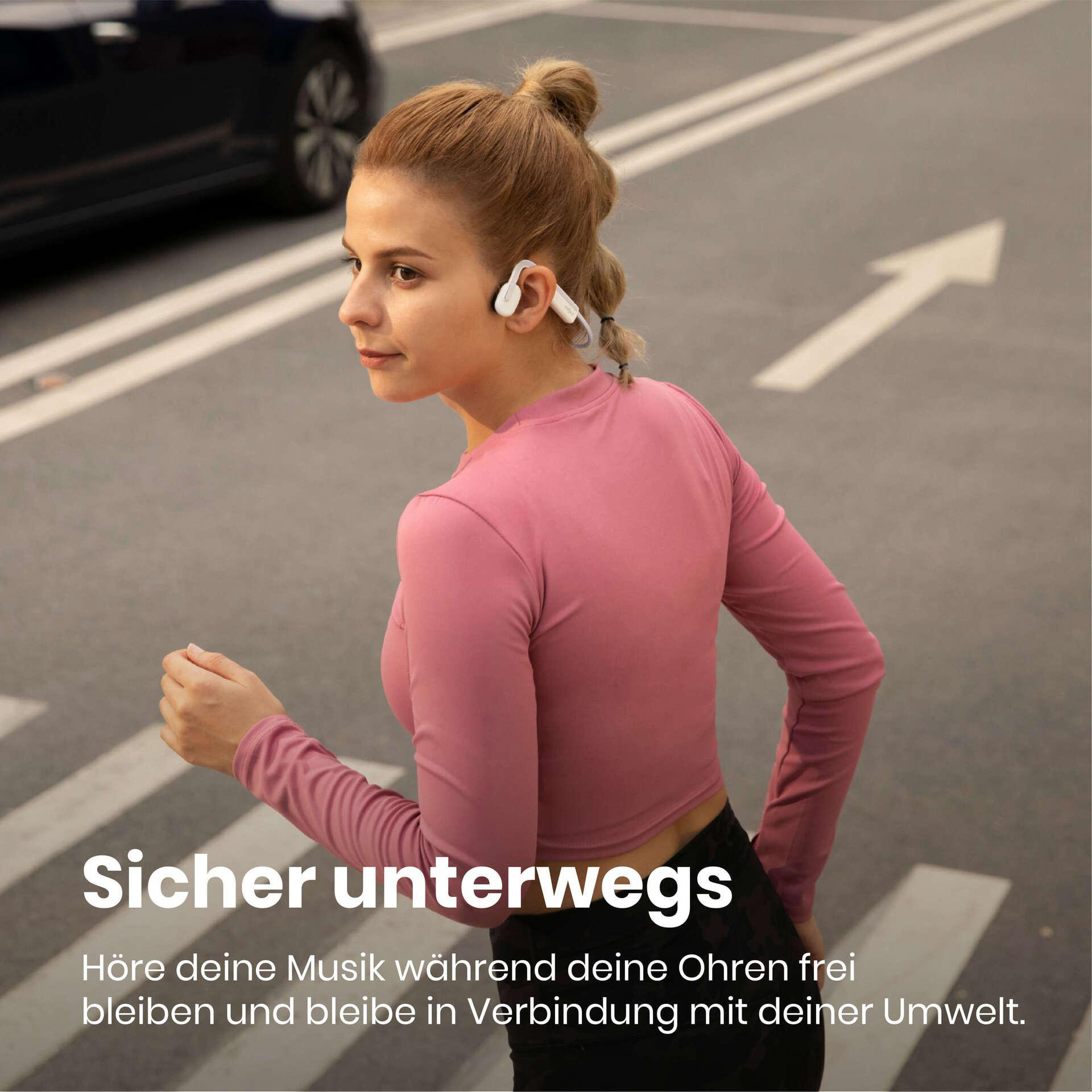 Eine Frau rennt auf einem Zebrastreifen und trägt ein rosa Hemd und weiße Kopfhörer. Ein Auto ist hinter ihr. Text: Sicher unterwegs.