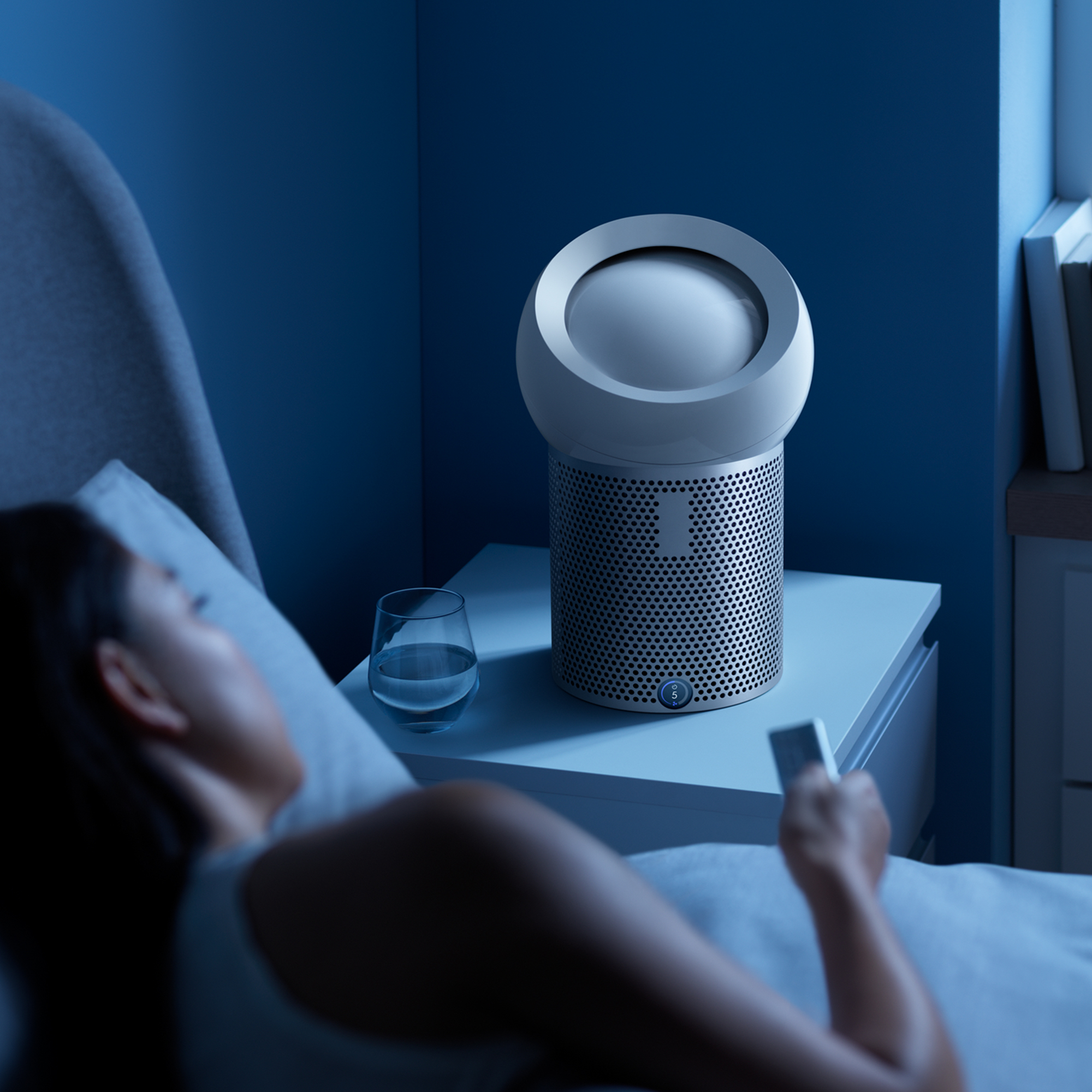 Una mujer en la cama con un ventilador en una mesita de noche, con un vaso de agua.